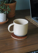 White mug on a leather coaster with a laptop and plants in the background