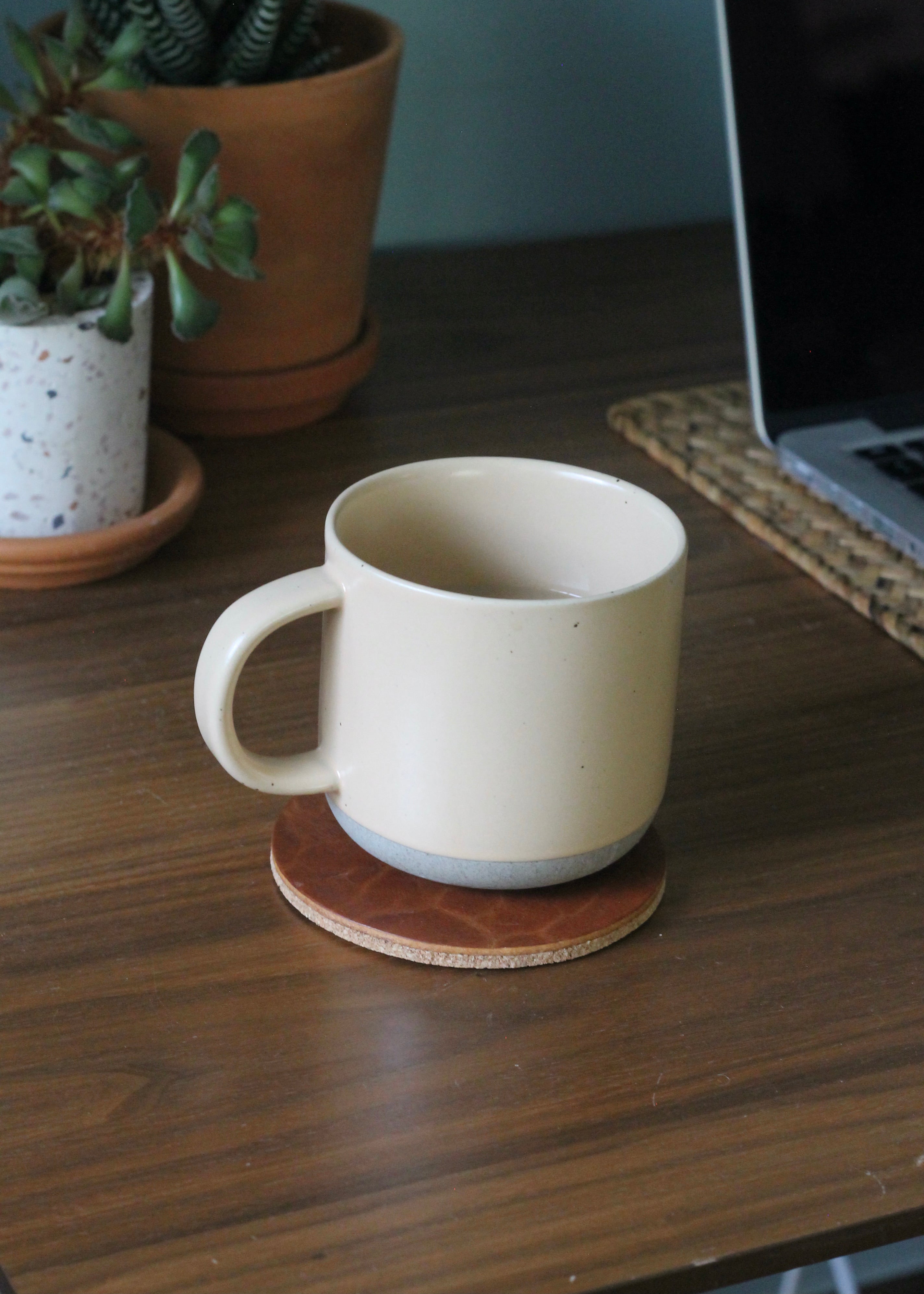 White mug on a leather coaster with a laptop and plants in the background